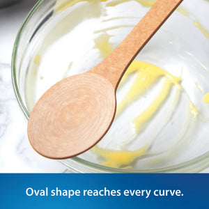 Close-up of the Everyday Stirring Spoon from Rada resting on top of a clear glass bowl with yellow cream or batter visible, on a white marble-like surface. A blue text banner at the bottom reads, 'Oval shape reaches every curve.