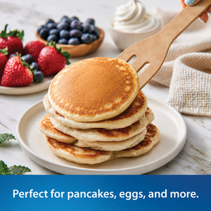 A stack of golden-brown pancakes on a white plate, being lifted with the Everyday Slotted Turner from Rada Cutlery. Fresh strawberries, blueberries, whipped cream, and a beige cloth are arranged on a light marble surface.