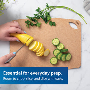 A person is cutting zucchini on the beige Essential Prep Board. The board is placed on a white marble countertop with sliced cucumbers and parsley nearby.