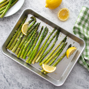 Roasted green asparagus spears on the Quarter Sheet baking pan with lemon slices, garnished with cheese, photographed from above on a light stone surface with lemons and a checkered cloth nearby.