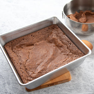 The Square metal baking pan from Rada shows baked chocolate brownies on a gray surface with a bowl of cocoa powder and an egg.