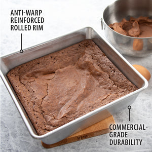 Rada's Square Pan containing a brown baked cake, shown on a light stone surface with a bowl of frosting and an egg in the background. Text callouts read 'ANTI-WARP REINFORCED ROLLED RIM' and 'COMMERCIAL-GRADE DURABILITY.