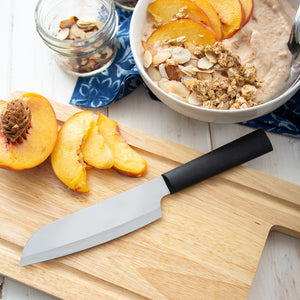 Black handled Cook's Utility Knife laying on a cutting board next to a sliced peach and a bowl of oatmeal.