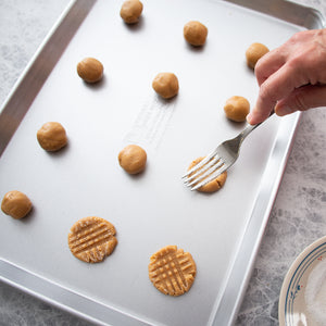 A hand with a fork flattening peanut butter cookie dough balls into criss-cross pattern.