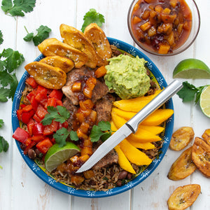 A silver Rada Cutlery serrated steak knife on a plate of carne asada beef with a side of mango, guacamole and plantains.