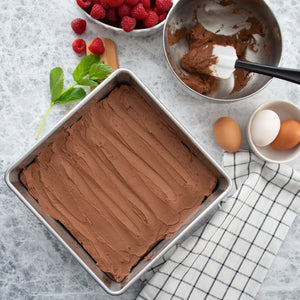 A pan of chocolate frosted brownies next to a bowl of frosting, a spatula, and a bowl of berries.