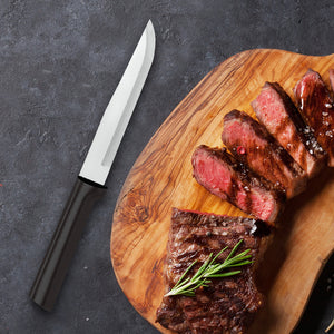 Rada's black handle Stubby Butcher next to a brown cutting board with a half sliced steak, with seasonings, greenery, and steak sauce on it.