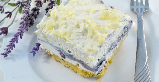 A colorful slice of Blueberry Lasagna with a graham cracker crust, blueberry middle and whipped topping with a fork to the right and some lavender laying to the left of the slice of dessert