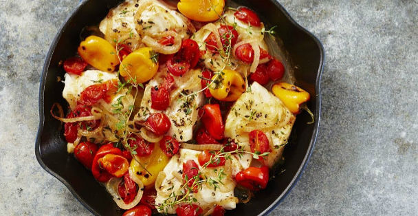 A black pan of Seared Cod with a buttery herb sauce and a mix of vegetables including sliced red and yellow peppers, tomatoes and onion on top of a granite countertop