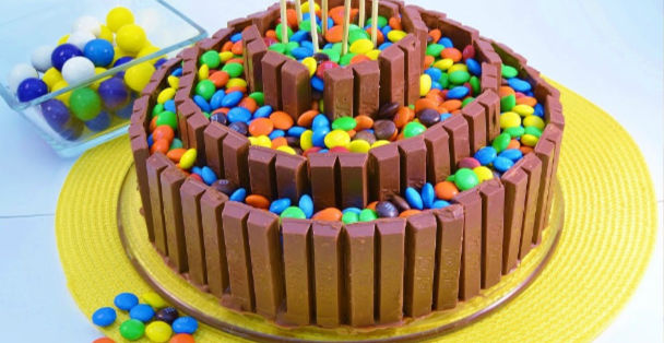 A three-ringed kit-kit bar cake with each ring full of multi-colored m&m's and candy pops on top of the cake atop a yellow sonnet placeholder next to a platter of candy pops 