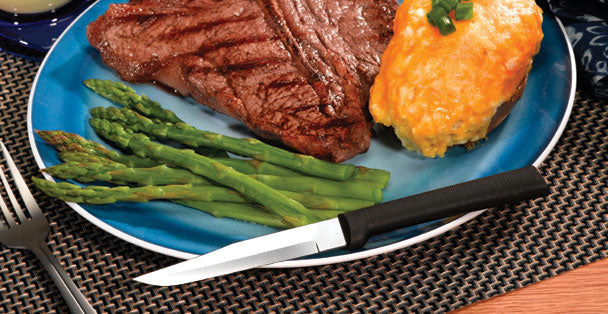 A RADA Cutlery Serrated Steak Knife with a black resin handle on a plate full of delicious asparagus, steak and cheesy potatoes