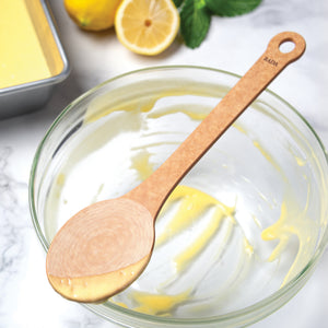 A wooden Everyday Stirring Spoon from Rada rests on top of a clear glass mixing bowl containing pale yellow batter or cream. The spoon has the brand name 'RADA' printed near the handle.