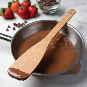 A Heavy-Duty Sauté Spoon rests on top of a stainless steel saucepan filled with melted chocolate. Fresh strawberries, loose chocolate chips, and a small bowl of chocolate chips are arranged on a light marble surface in the background.