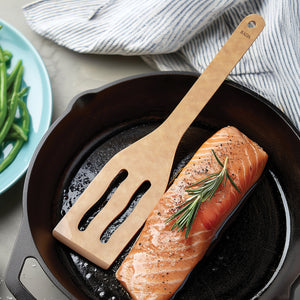 Rada's wooden Everyday Slotted Turner resting on a black cast iron skillet next to a cooked salmon fillet with a rosemary sprig. A light blue plate with green beans is partially visible on the left, and a striped kitchen towel is in the background.
