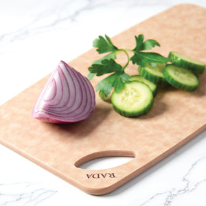 A close-up of Rada's Small Prep Board showing the built-in hanging hole with 'RADA' engraved in dark brown. A red onion wedge and sliced cucumber with a sprig of basil sit on top of the cutting board on a white marble surface.