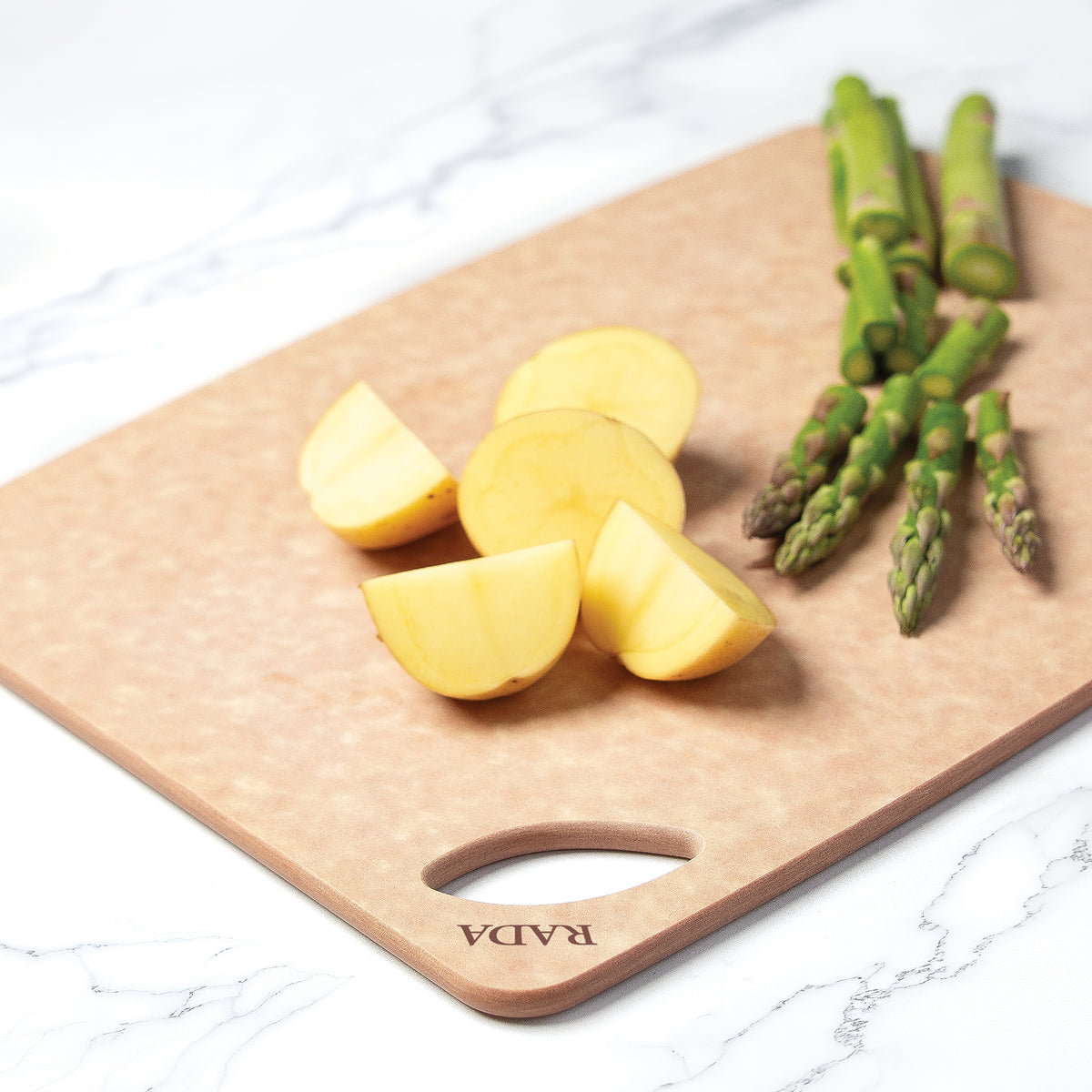 Rada's Essential Prep wooden cutting board with a handle cutout, featuring the brand name 'RADA' printed in the corner. The board is styled with sliced yellow potatoes and green asparagus on a white marble surface