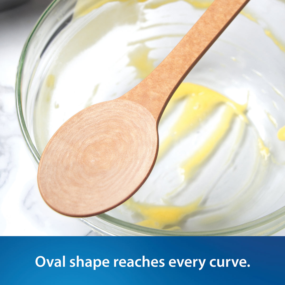 Close-up of the Everyday Stirring Spoon from Rada resting on top of a clear glass bowl with yellow cream or batter visible, on a white marble-like surface. A blue text banner at the bottom reads, 'Oval shape reaches every curve.