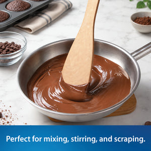 A stainless steel saucepan filled with smooth melted chocolate, with Rada's Heavy Duty Sauté Spoon stirring inside. The pan sits on a light marble surface with baking ingredients and a muffin tray in the background.