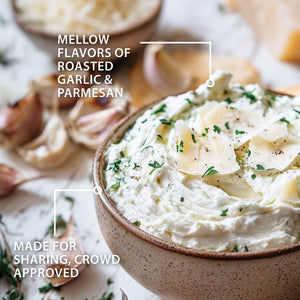 A close-up of the Roasted Garlic and Parmesan Dip in a speckled brown ceramic bowl, garnished with fresh parsley and shaved cheese. Blurred garlic cloves and herbs sit in the background.