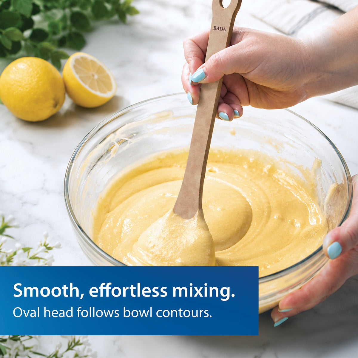 A person using the Everyday Stirring Spoon to mix pale yellow batter in a glass bowl. Lemons and herbs sit in the background on a white marble surface, with a blue text banner reading 'Smooth, effortless mixing. Oval head follows bowl contours.'