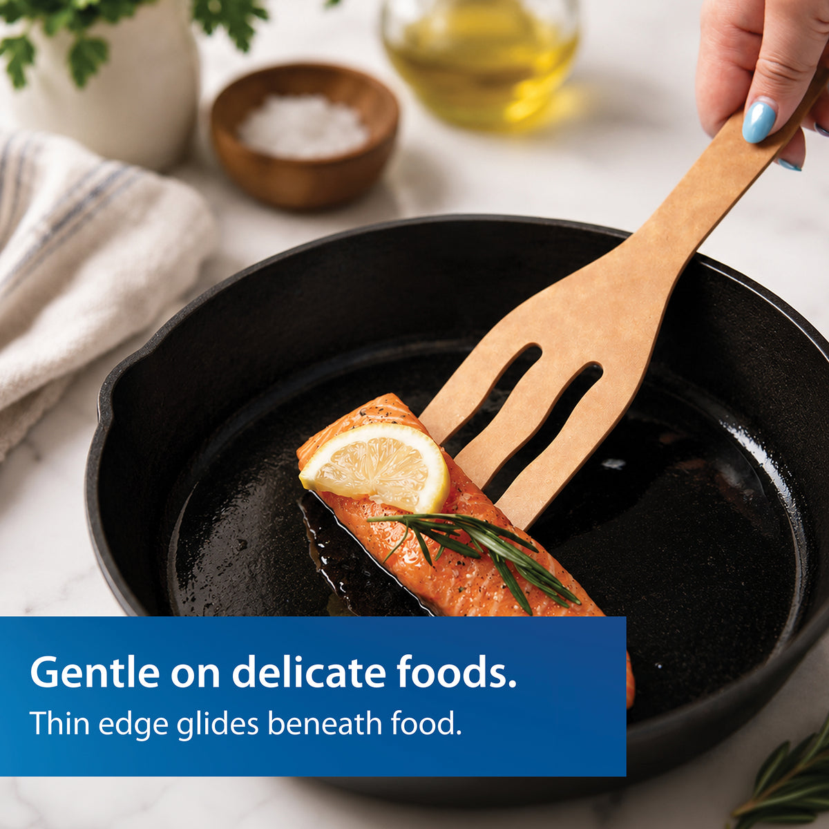 The Everyday Slotted Turned from Rada is shown lifting salmon from a black cast iron skillet on a white marble countertop. A blue text banner at the bottom reads, 'Gentle on delicate foods. Thin edge glides beneath food.'