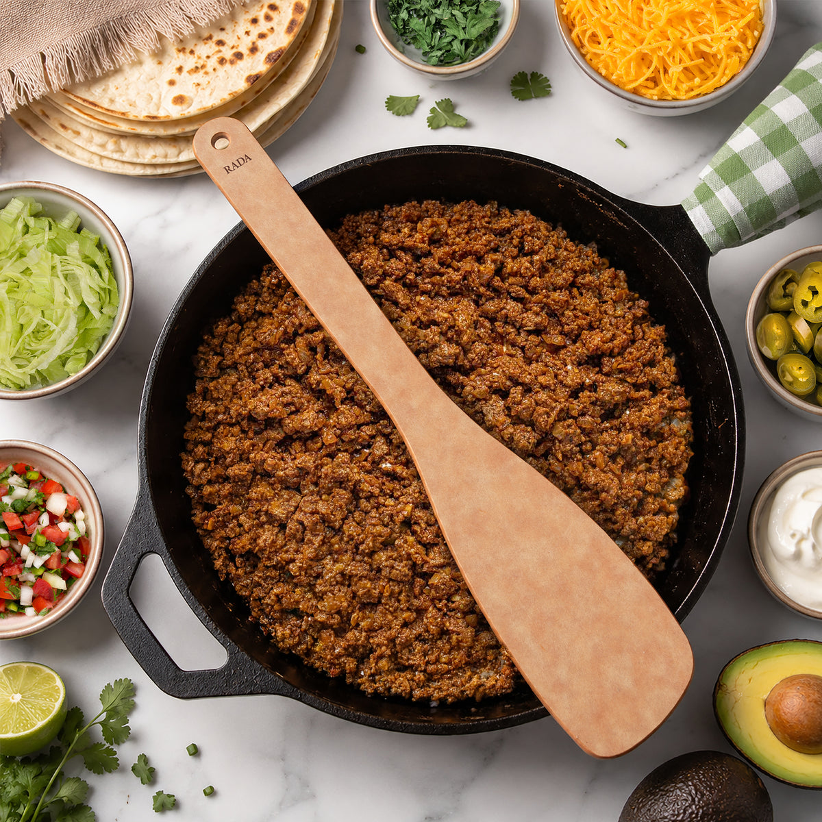 A Heavy-Duty Saute Spoon sits on a black cast iron skillet filled with cooked ground meat. The skillet is surrounded by bowls of taco ingredients including shredded lettuce, shredded cheese, olives, sour cream, cilantro, lime, and tortillas.