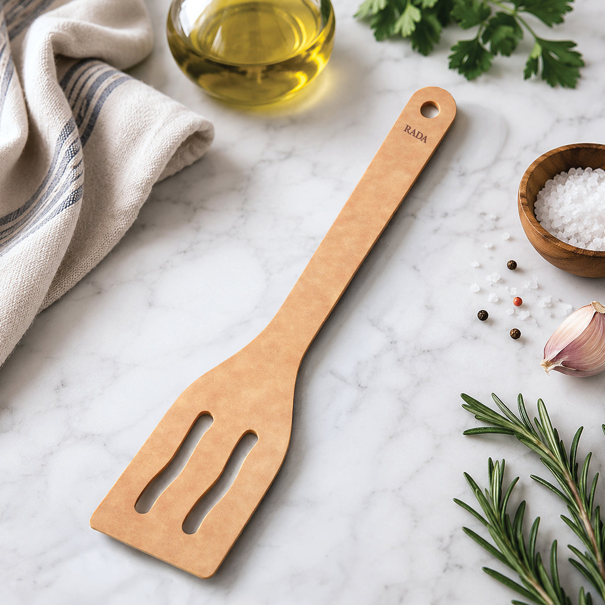 Rada Cutlery's Everyday Slotted Turner engraved with a handle label reading 'RADA' sits on a white marble surface next to rosemary, a sprig of basil, and small bowls of olive oil and sea salt.