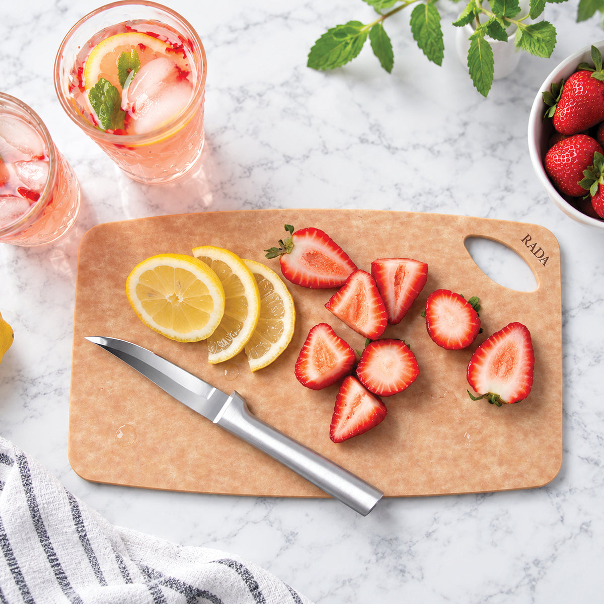 The wooden Small Prep cutting board with a silver knife, styled with sliced strawberries, lemon slices, and glasses of pink drink on a white marble surface.