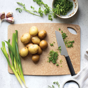 Rada's Essential Prep cutting board shown in a styled kitchen flat-lay with potatoes, green onions, garlic, parsley, a knife, and a bowl. The cutting board has a subtle embossed pattern and the brand name 'RADA' is engraved on the top right corner.