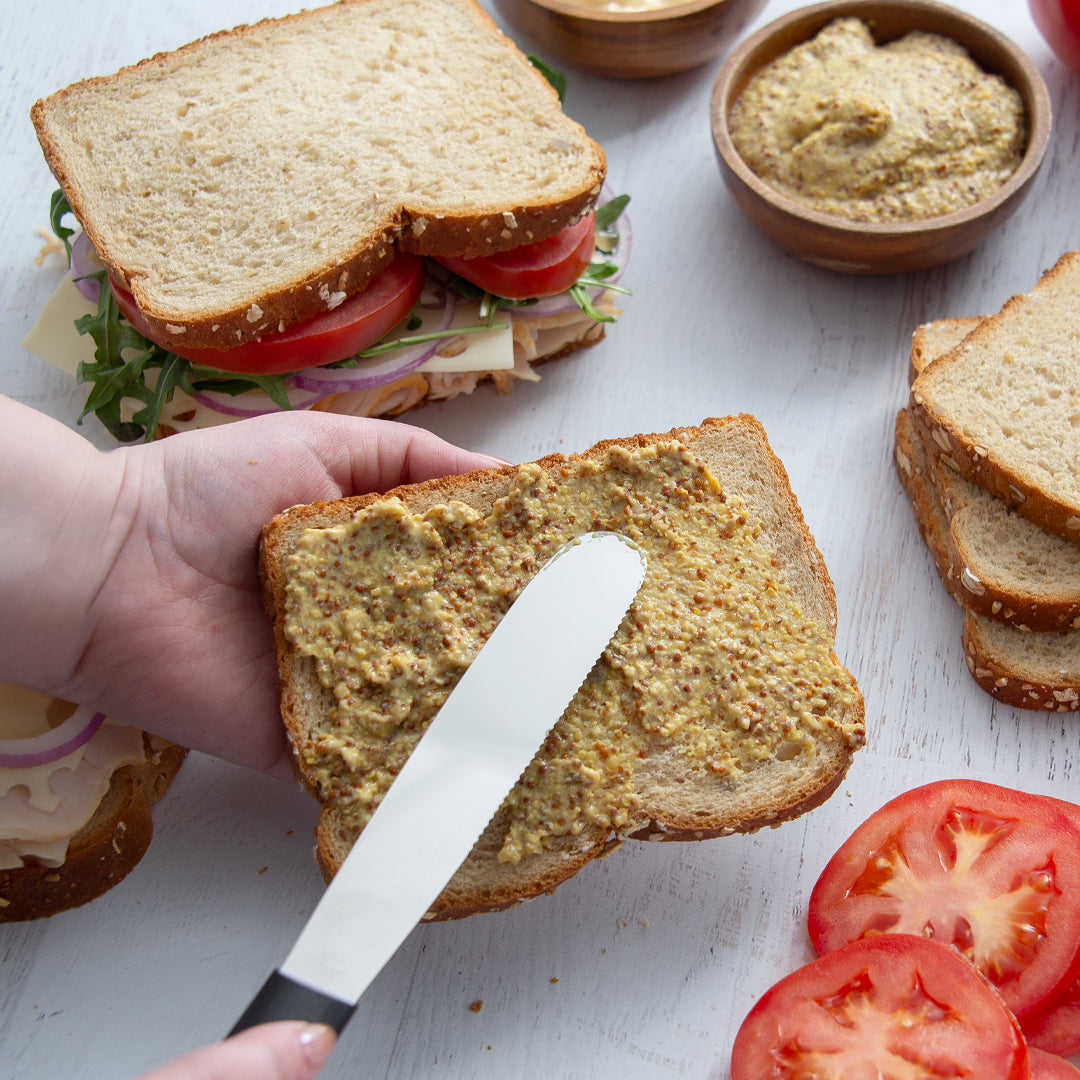 Person spreading mustard on a slice of bread with a sandwich in the background.