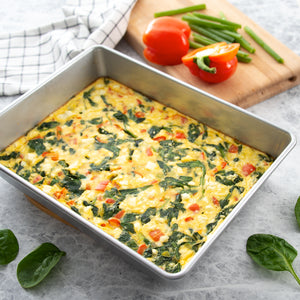 A Cake Pan from Rada Cutlery used to make a baked spinach and tomato frittata, styled on a light gray surface with fresh spinach leaves, a cutting board with vegetables, and a checkered cloth.