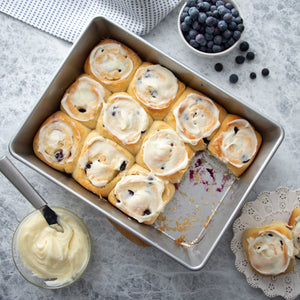 A rectangular metal Cake Pan from Rada Cutlery filled with freshly baked blueberry cinnamon rolls topped with white frosting, with one roll cut out. A small bowl of blueberries, a glass of frosting, and a cloth napkin are arranged around the pan on a light gray surface.