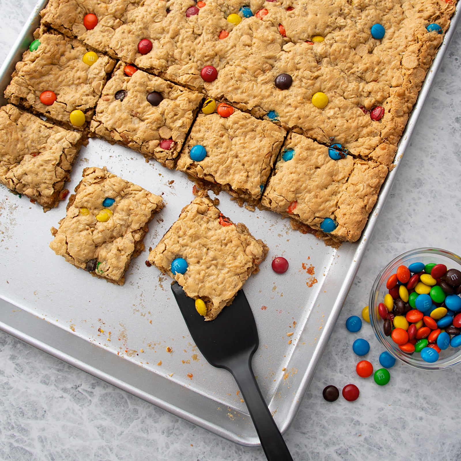 Rada's Half Sheet Pan with caramel oatmeal cookie bars topped with colorful M&M's candies, arranged with a black spatula and a small glass bowl of candy-coated chocolate pieces on a light gray surface.