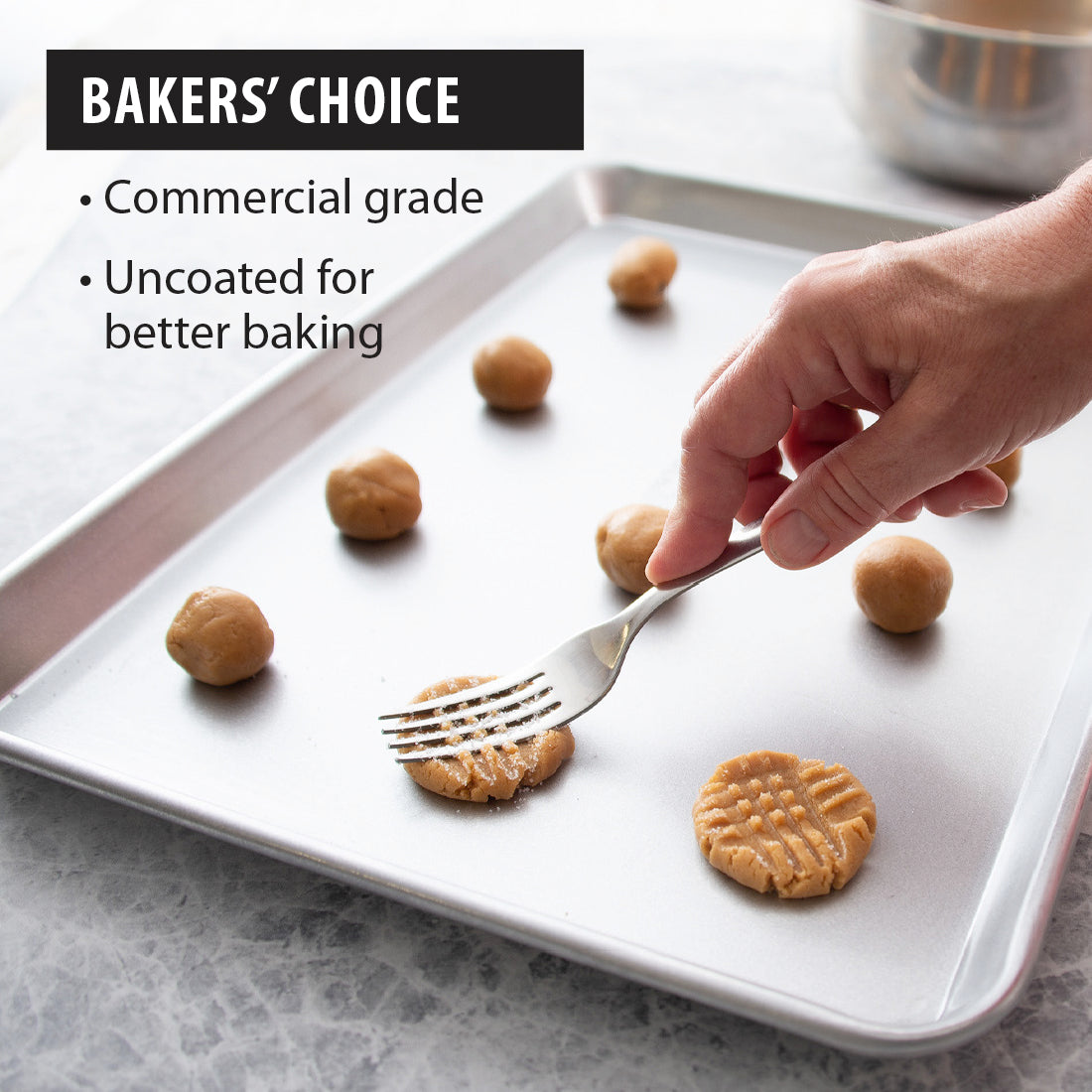 Hand using a fork to shape cookie dough balls on a Half Sheet Pan from Rada with callouts highlighting its commercial grade and uncoated design for better baking.