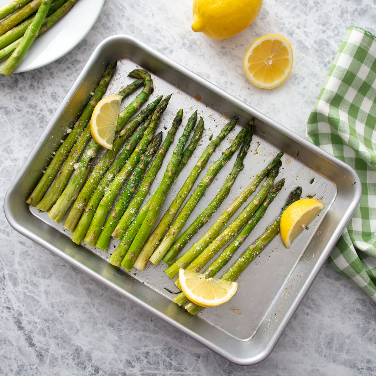 Roasted green asparagus spears on the Quarter Sheet baking pan with lemon slices, garnished with cheese, photographed from above on a light stone surface with lemons and a checkered cloth nearby.