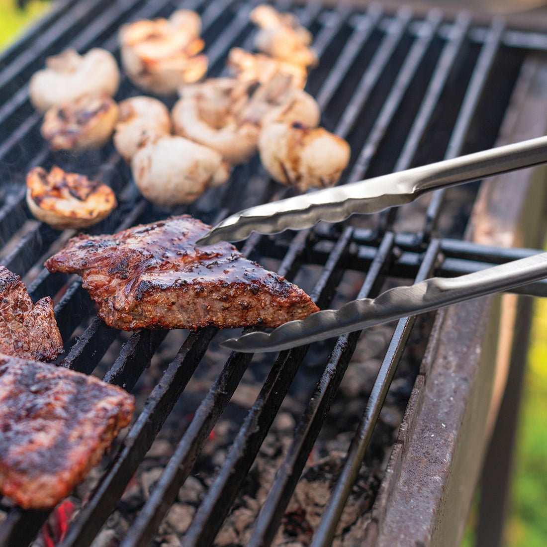 12 inch tongs on the grill with meat and mushrooms, flipping the meat on the grill.