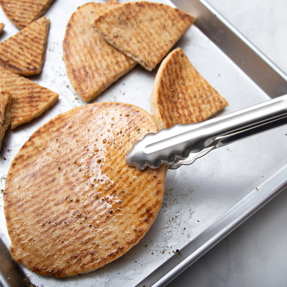 9 inch Tongs picking up homemade pita chips off a half sheet pan while cooking.