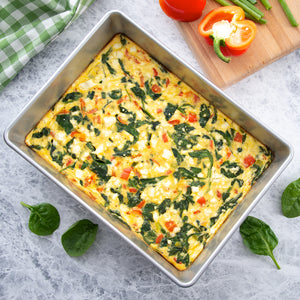 Spinach, feta and tomato egg casserole ready to be cut surrounded by fresh spinach leaves.
