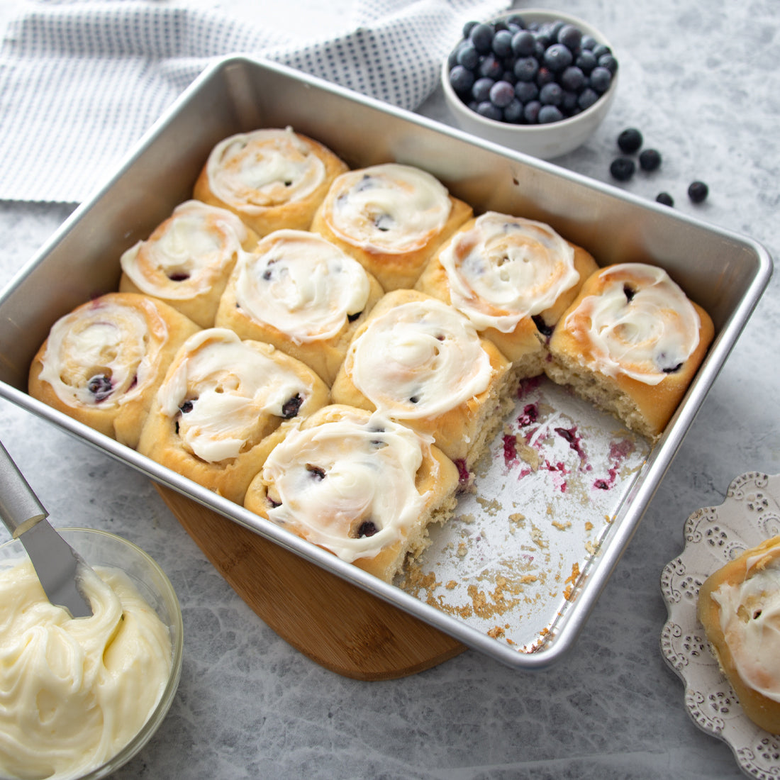 Cake pan with blueberry sweet rolls and cream cheese frosting alongside a bowl of fresh blueberries.