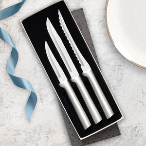 A gift boxed set of three kitchen knives, a paring, utility/steak and tomato slicer laying beside a plate with a blue ribbon.