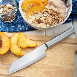 Sliced peaches on a cutting board with a Cook's Utility knife next to a bowl of oatmeal with slivered almonds and granola.