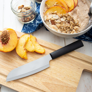 Black handled Cook's Utility Knife laying on a cutting board next to a sliced peach and a bowl of oatmeal. 