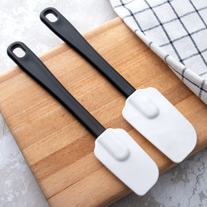 Two flexible head spatulas with black handles laying on a wood cutting board. Sturdy handles with a hole for hanging. 