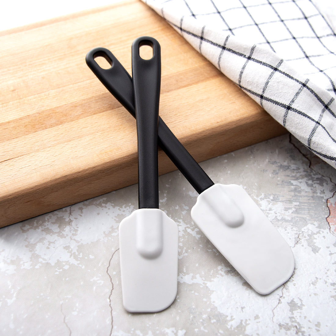 Two flexible Rada Cutlery spatulas leaning on a wood cutting board with a checkered dish towel. 