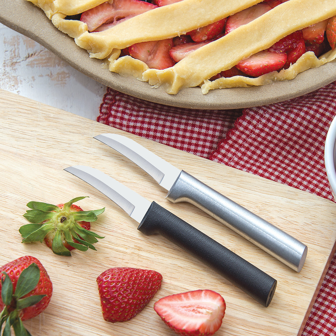Two Granny paring knives laying on a cutting board with sliced strawberries next to a unbaked strawberry pie. 