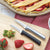 Two Granny paring knives laying on a cutting board with sliced strawberries next to a unbaked strawberry pie. 