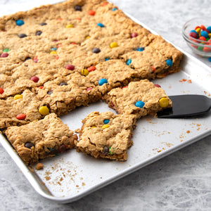 Half sheet pan with monster cookie bars on gray countertop. Spatula under bars ready to serve.
