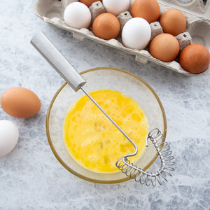 Handi-Stir whisk with silver handle on a bowl of beaten eggs with brown and white eggs in background