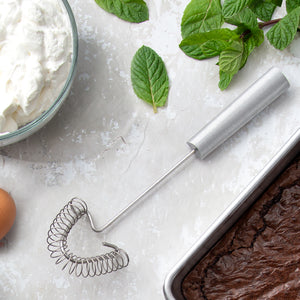 Rada's silver handle Handi-Stir on a light background next to a pan of brownies, leafy greens, and a bowl of frosting.