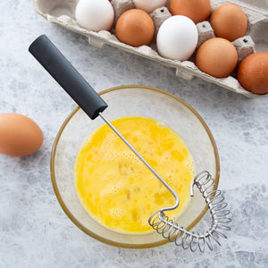 Black handled Handi-Stir on a bowl of whisked eggs. Carton full of eggs sitting on a gray countertop.